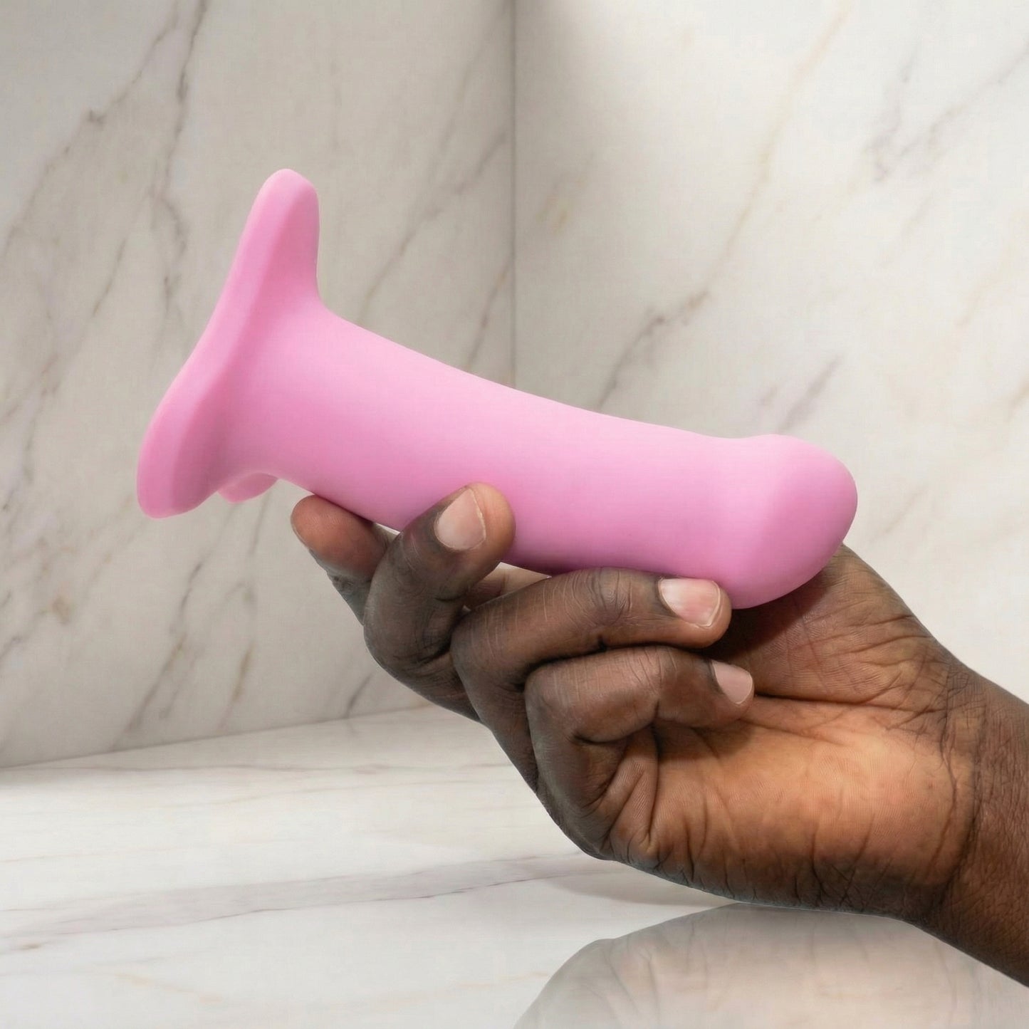 Roze siliconen dildo met zuignap basis, vastgehouden in hand tegen marmeren achtergrond