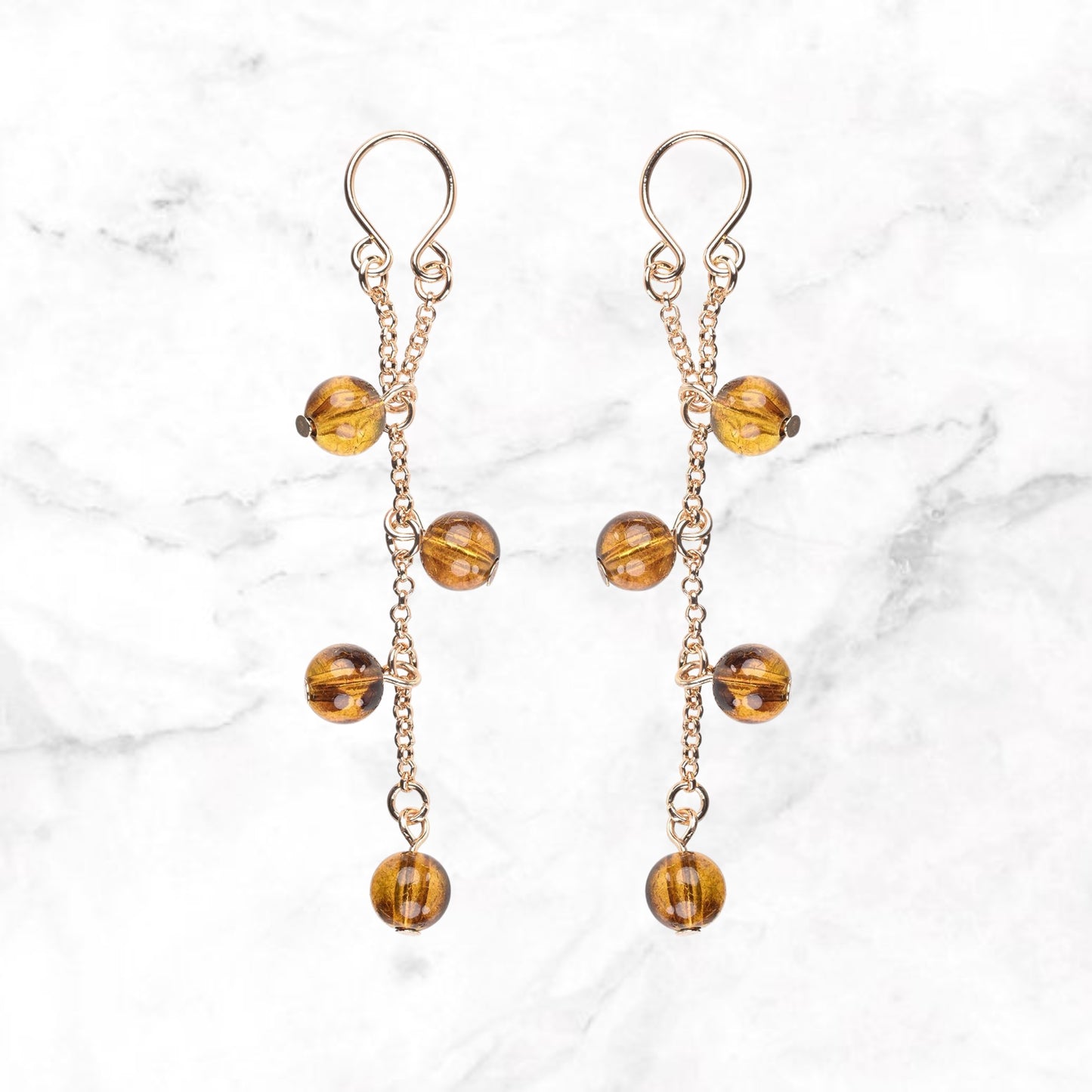 Twee tepelsieraden met goudkleurige kettinkjes en amberkleurige bolkralen op marmer